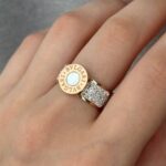 Широка каблучка BVLGARI з круглим логотипом та камінням золото білий на руці